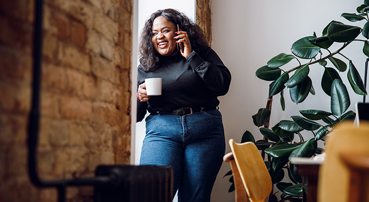 Positive Afro American lady looking out of the window while talking on the phone and drinking tea