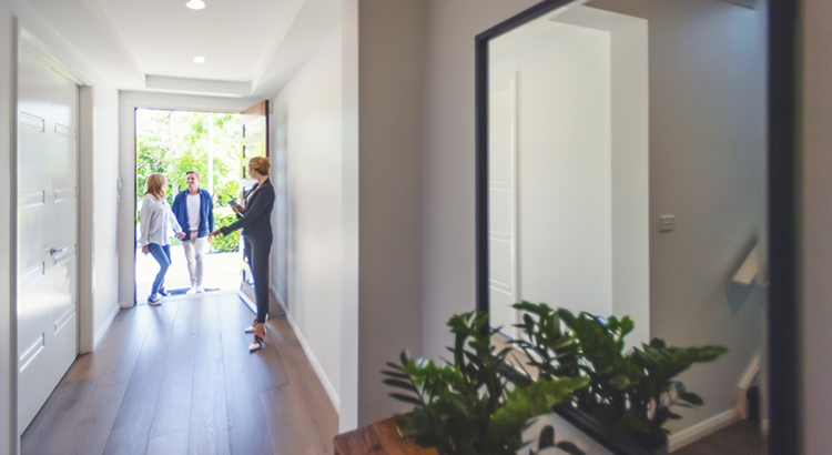 Real estate agent showing a young couple a new house. The house is contemporary. All are happy and smiling. The couple are casually dressed and the agent is in a suit. The couple are being greeted at the front door by the real estate agent. Copy space