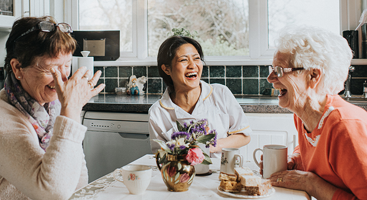 A female care assistant visits two older ladies in their home during a home visit. They sit at a kitchen table and drink tea while laughing together. They are all very comfortable and happy as they gesture and laugh loudly. The scene is upbeat and fun. Widow provides a space for copy.
