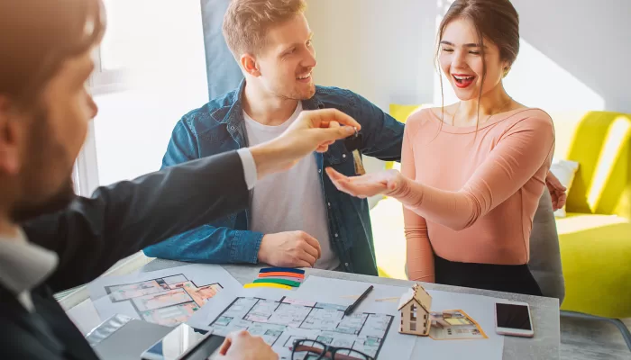 Couple buy or rent apartment together. Happy young woman reach hand and get keys from realtor. Man look at her and smile. Bright sunlight from window